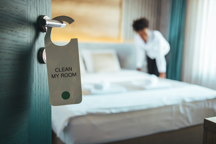 Türschild mit der Aufschrift „Clean My Room“ und Housekeeping-Mitarbeiterin bei der Zimmerkontrolle im Hintergrund – Glanz & Glorie Hotel Service